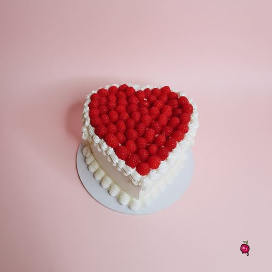 A heart-shaped cake decorated with a large red raspberry heart on top and white piped frosting borders, sitting on a white plate against a white marble-patterned background.