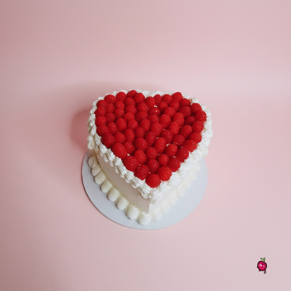 A heart-shaped cake decorated with a large red raspberry heart on top and white piped frosting borders, sitting on a white plate against a white marble-patterned background.