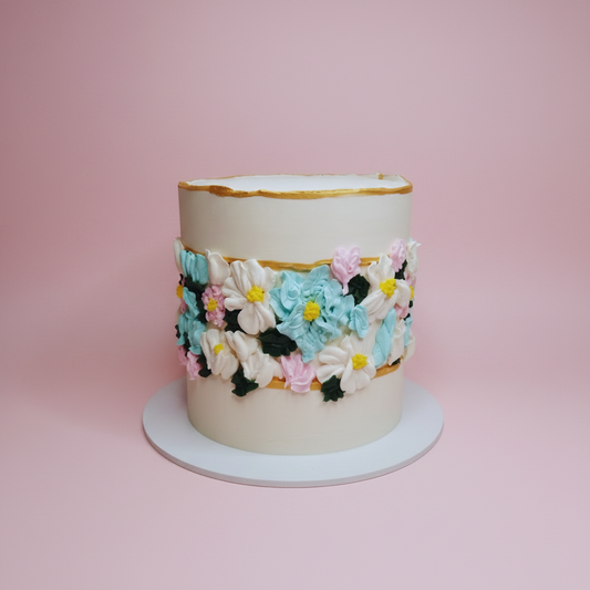 A cylindrical cake with off-white frosting and a decorative band of colorful piped flowers, set on a white round cake board against a white marble-patterned background.