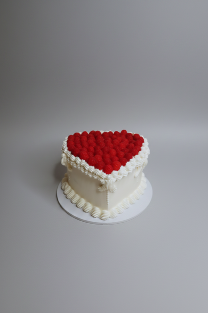 A heart-shaped white cake decorated with a red textured top and white piped borders, sitting on a white surface against a white marble-patterned background.